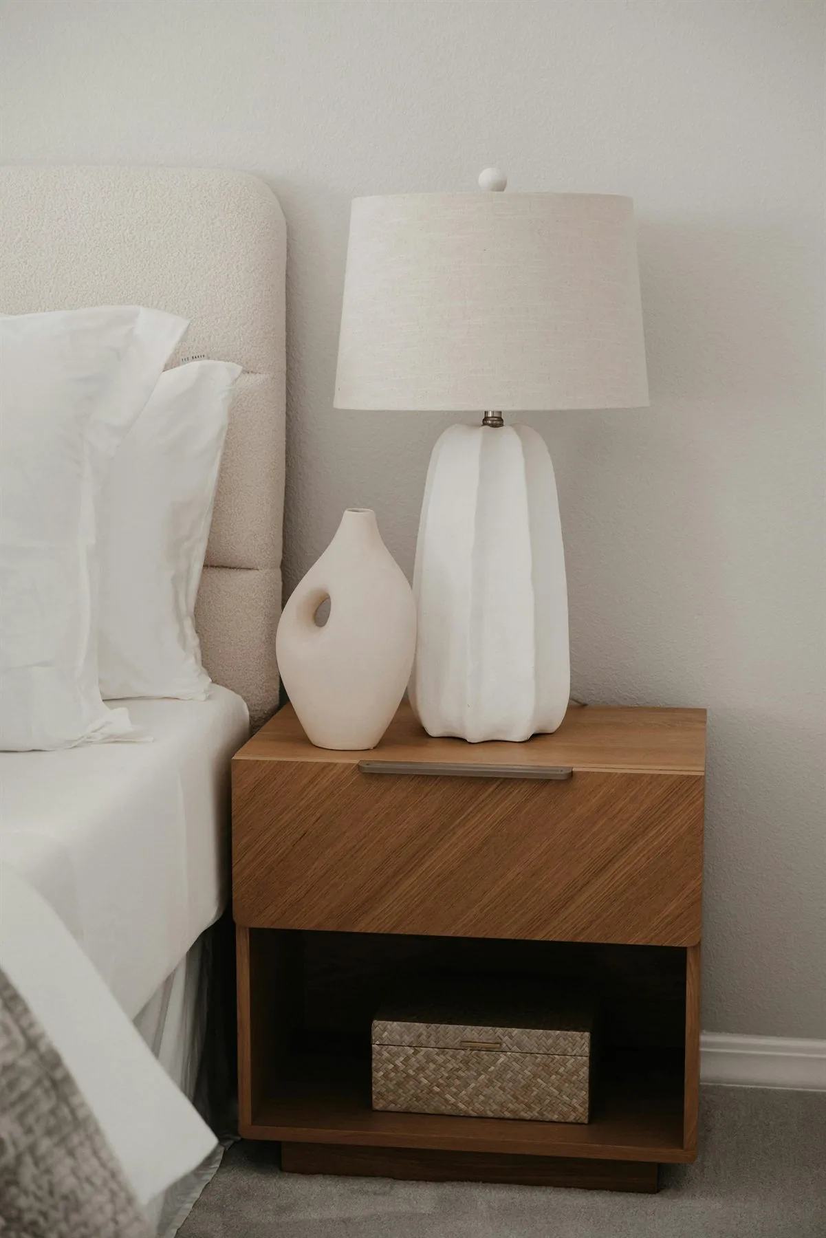 Minimal bedside table with a white lamp, wood drawer, and soft neutral bedding