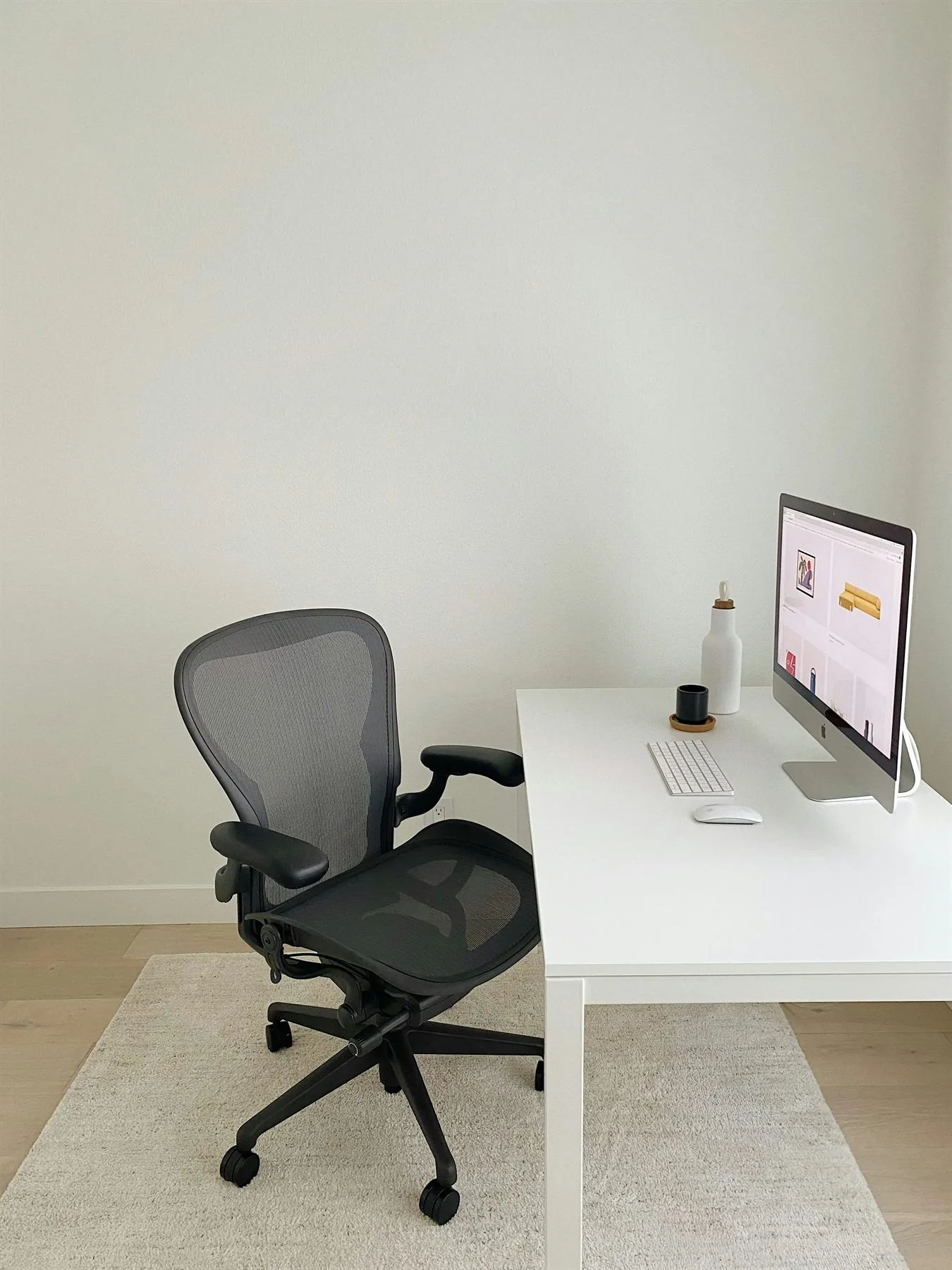 Minimal office with a white desk, black task chair, pale rug, and warm off-white wall