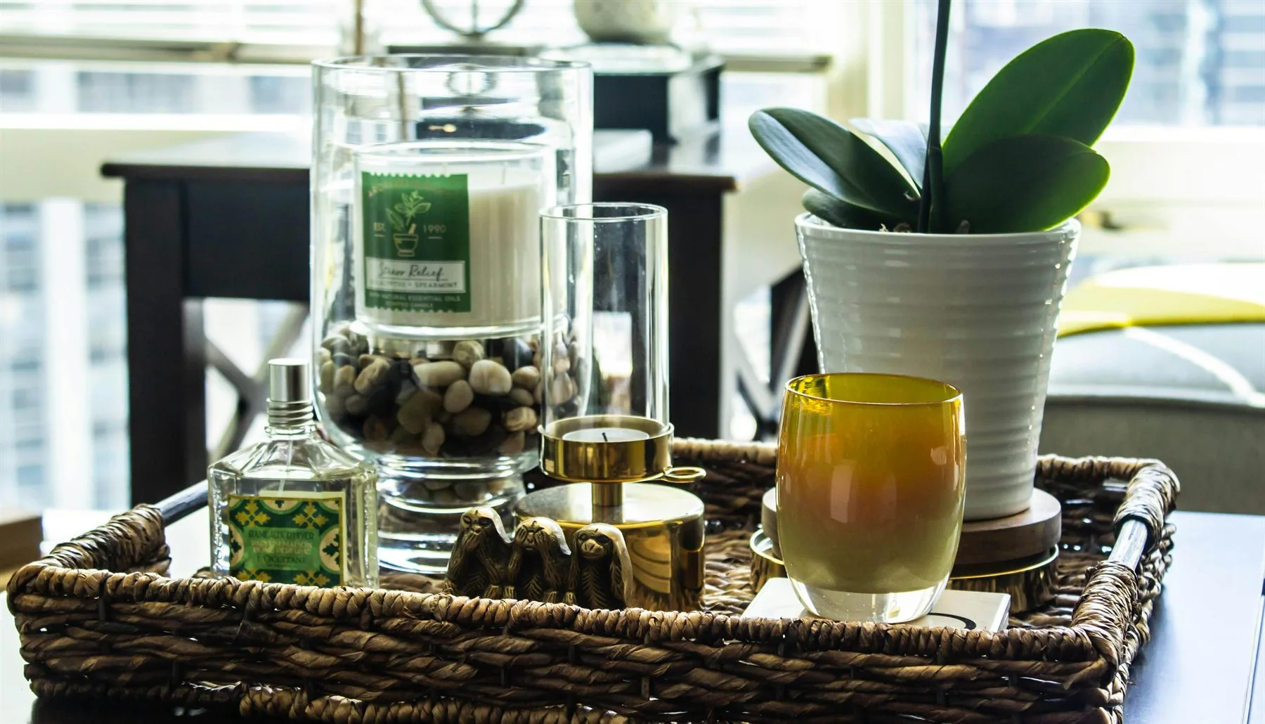 Woven tray with a plant, candle, glassware, and small decor pieces near a window